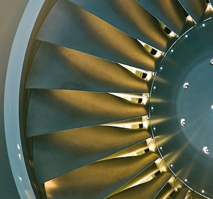 Close-up of an airplane engine turbine, showing the metallic fan blades arranged in a circular pattern. The blades reflect light, highlighting their sharp edges and mechanical precision. The image emphasizes the engineering and functionality of aircraft propulsion systems.